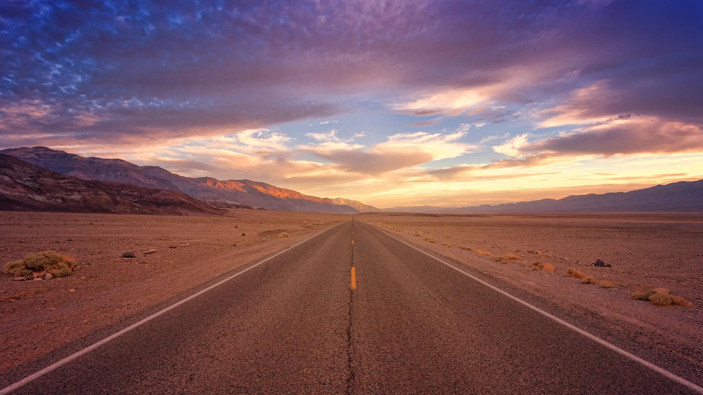 A long desert highway leading toward mountains beneath a colorful sunset